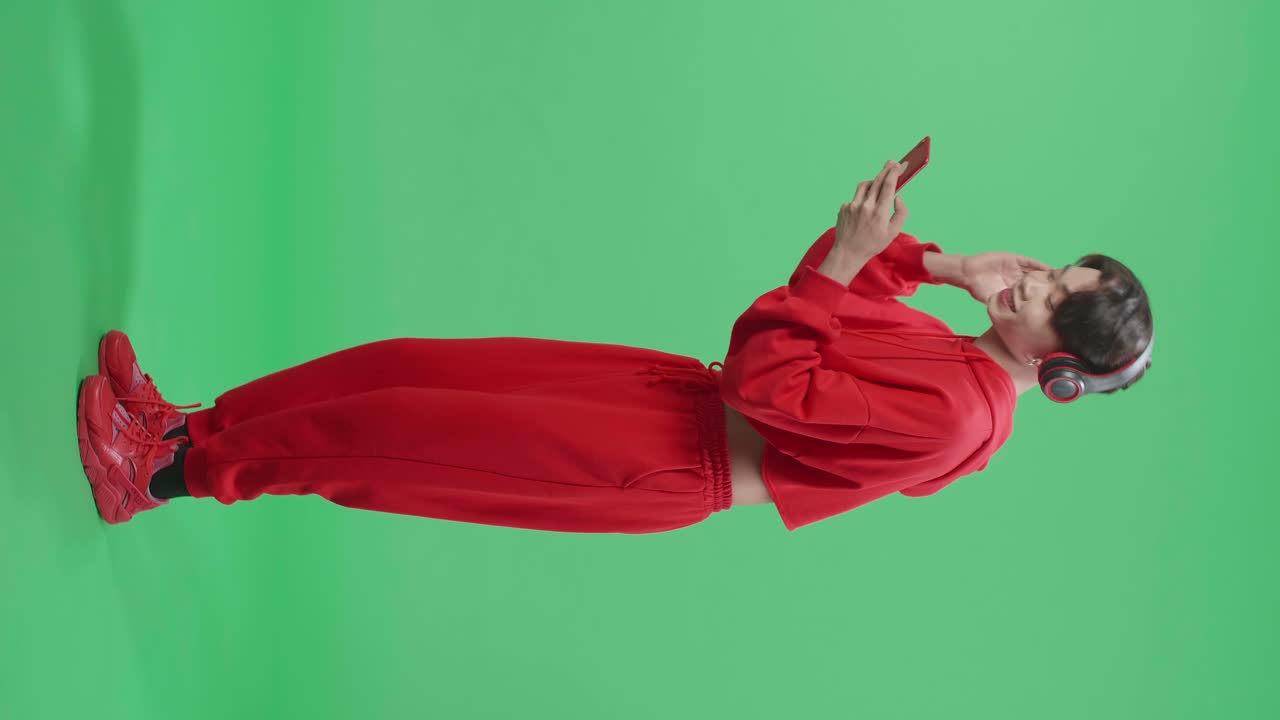 Full Body Of Side View Of Asian Transgender Male With Headphones Listening To Music From Mobile Phone And Dancing In The Green Screen Studio