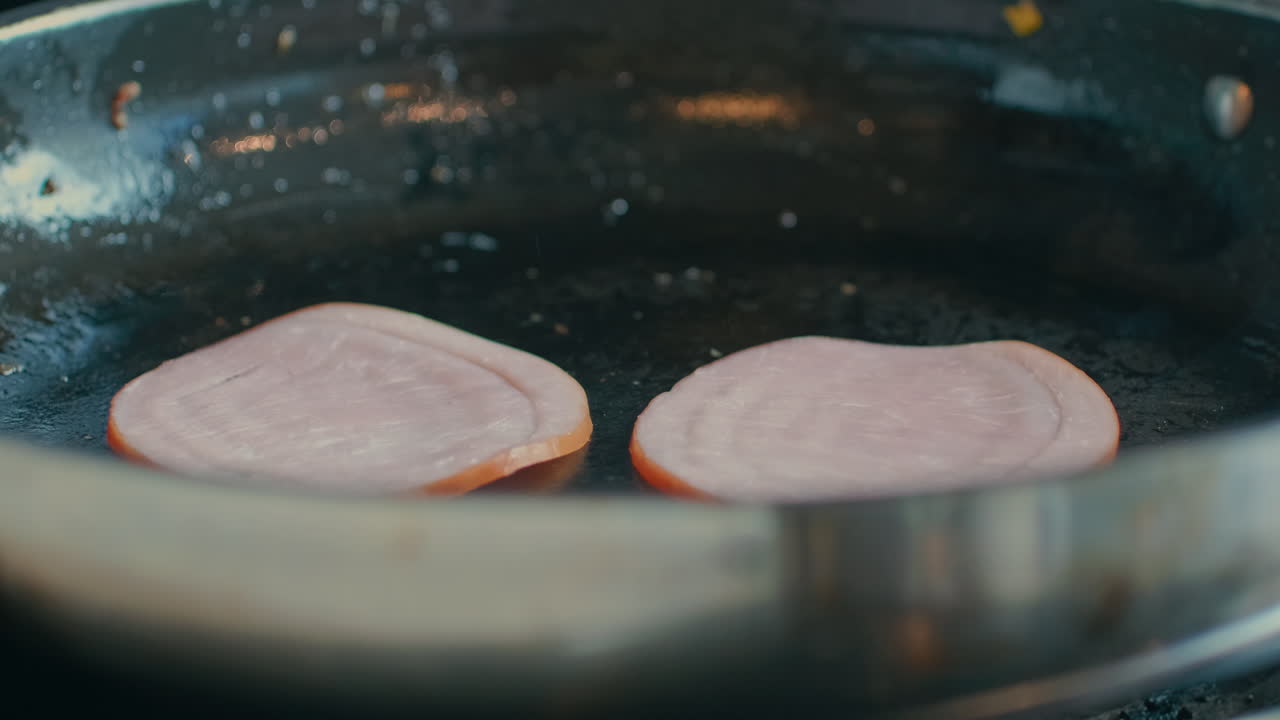 Close-up of a hand carefully placing two slices of Canadian bacon or ham onto a hot stainless steel skillet to begin frying for a breakfast meal