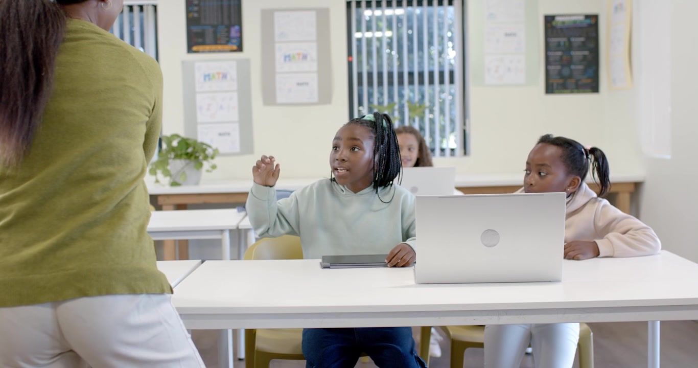 Insegnante afroamericana che gesticola, avvicinandosi e coinvolgendo giovani ragazze che usano un tablet in classe.