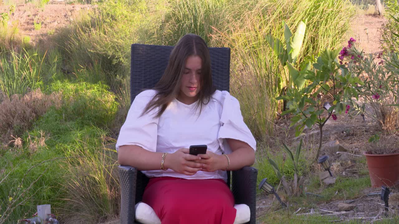 A young girl sits in a garden chair and types on her smartphone