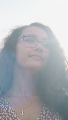 mujer con gafas y colgante de cruz ajusta su cabello al aire libre, sonriendo cálidamente mientras la luz del sol se refleja en su cara, rodeada de vegetación y una suave luz natural