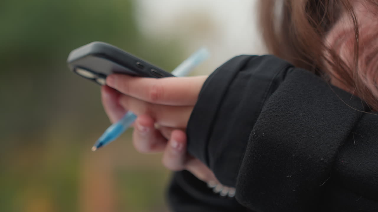 Studente scorre il telefono con una penna, primo piano laterale delle mani che tengono uno smartphone e una penna blu in una felpa con cappuccio nera, sfondo sfocato di un parco, multitasking tra messaggi e appunti veloci, spontaneo