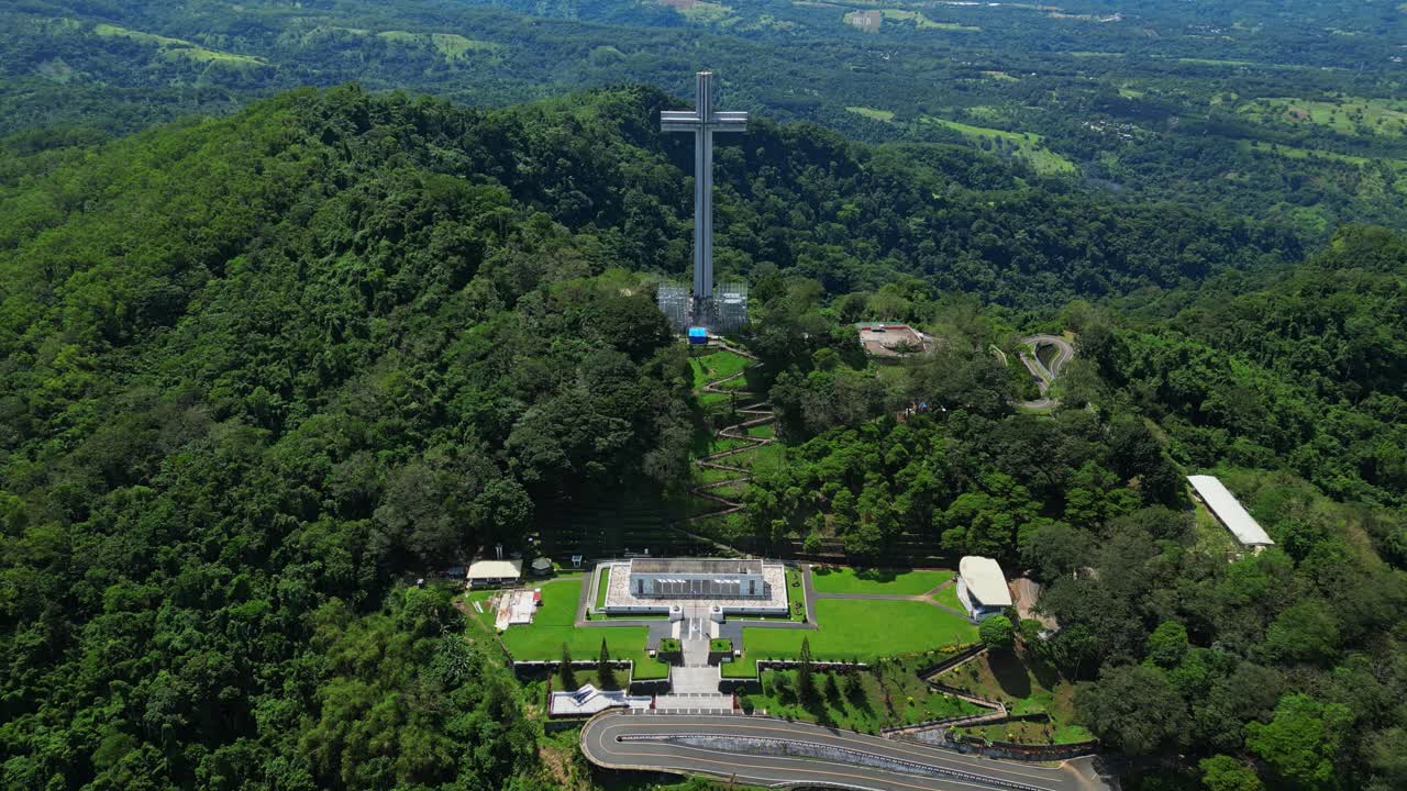 Front‑view push aerial highlighting the towering Mt. Samat National Shrine, revealing its monumental cross framed by landscaped grounds and lush forested hills in Pilar, Bataan, Philippines