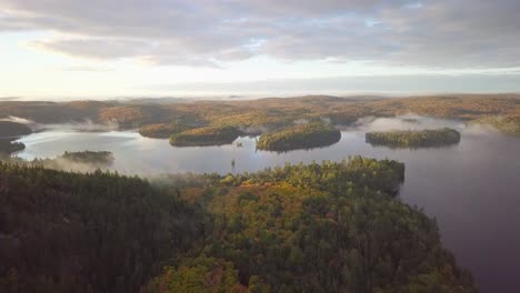 Toma-Amplia-Aérea-Del-Amanecer-Sobrevolando-Los-Colores-Del-Bosque-Otoñal-Hacia-Lagos-Brumosos-Con-Islas-Cubiertas-De-Niebla-En-Kawarthas,-Ontario,-Canadá