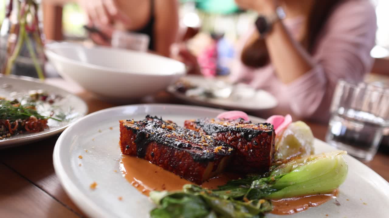 A close-up view of a gourmet dish featuring thick sliced bacon, greens, and sauce in a lively restaurant setting