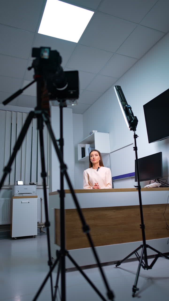 Long-haired woman in white jacket sits at desk indoors. Professional camera on tripod records a video with female blogger. Vertical video.