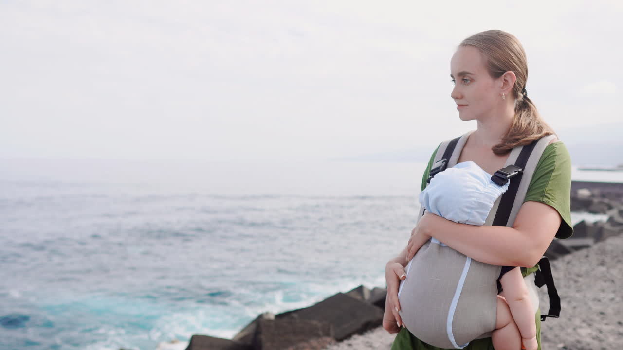 Along the coastline of the ocean, a mother and her baby take a leisurely stroll on a sunny summer day, the child secured in a kangaroo backpack