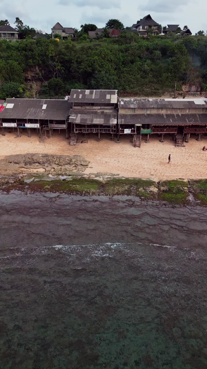 Vertical aerial shot of a coastal village with simple wooden cabins and beach huts built near the sand overlooking reef covered shallows and calm tropical waters typical of Southeast Asian shores
