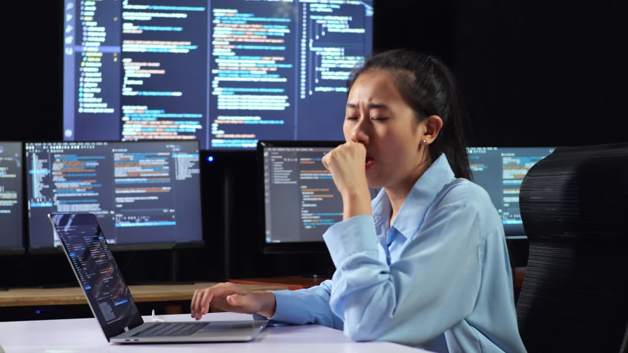 Side View Of Asian Female Programmer Yawning And Sleeping While Writing Code By A Laptop Using Multiple Monitors Showing Database On Terminal Window Desktops In The Office