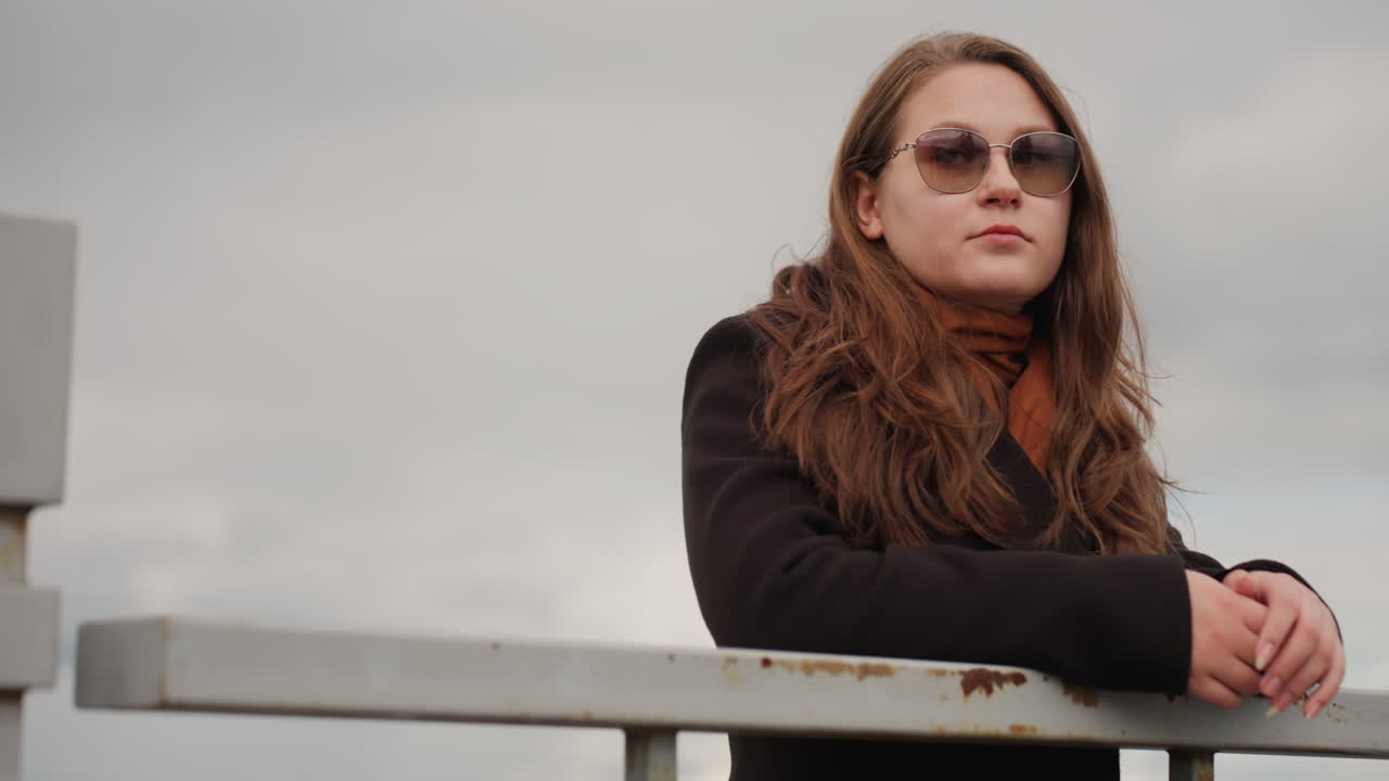 Fair skinned girl with blonde hair leans on iron rail while looking forward under cloudy sky, expressing calm determination, peaceful reflection, and quiet confidence in natural outdoor setting