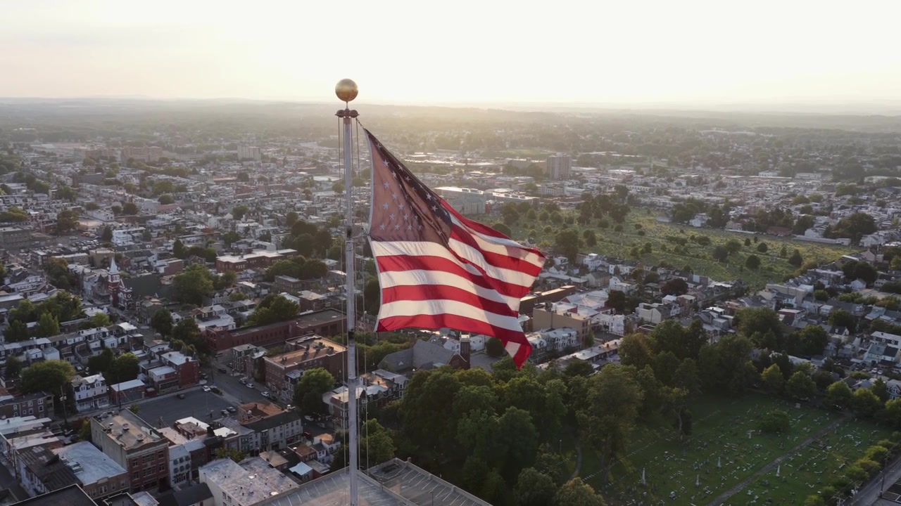 Gran bandera estadounidense ondea en lo alto de un mástil sobre el centro de Allentown, Pensilvania, con densos tejados y calles residenciales arboladas que se extienden por el ajetreado paisaje urbano de abajo. Toma orbital de dron aéreo
