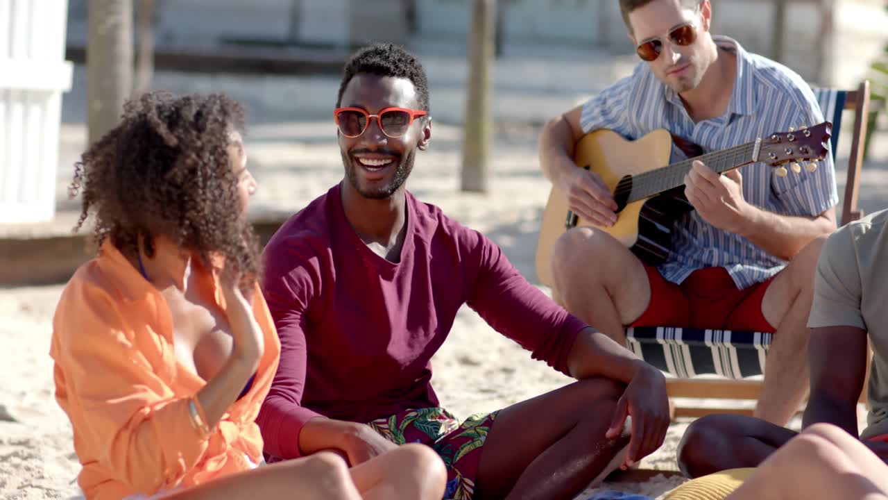 Felice gruppo eterogeneo di amici seduto che suona la chitarra sulla spiaggia con palme e una casa al mare