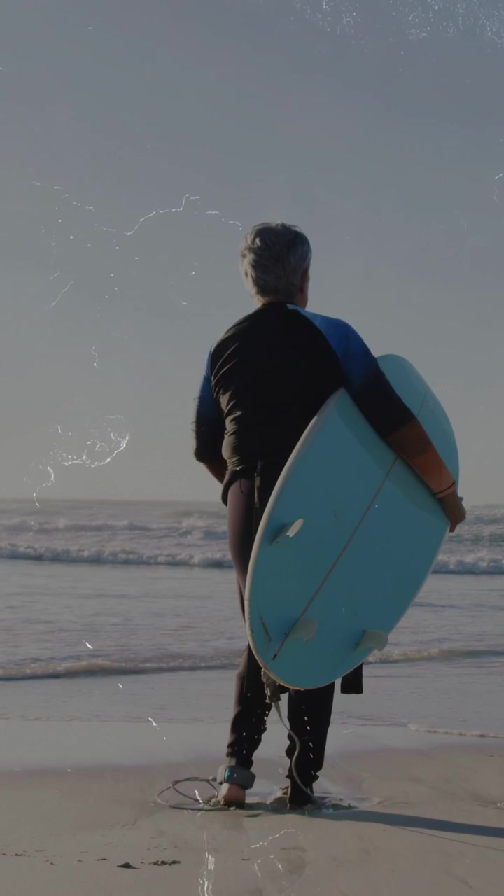 Video verticale: Surfista anziano che tiene la tavola, dopo aver cambiato la presa, camminando verso le onde per fare sport.