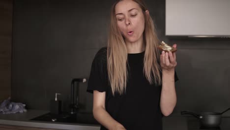 blonde woman eating bread with butter for breakfast, checking her smartphone in kitchen