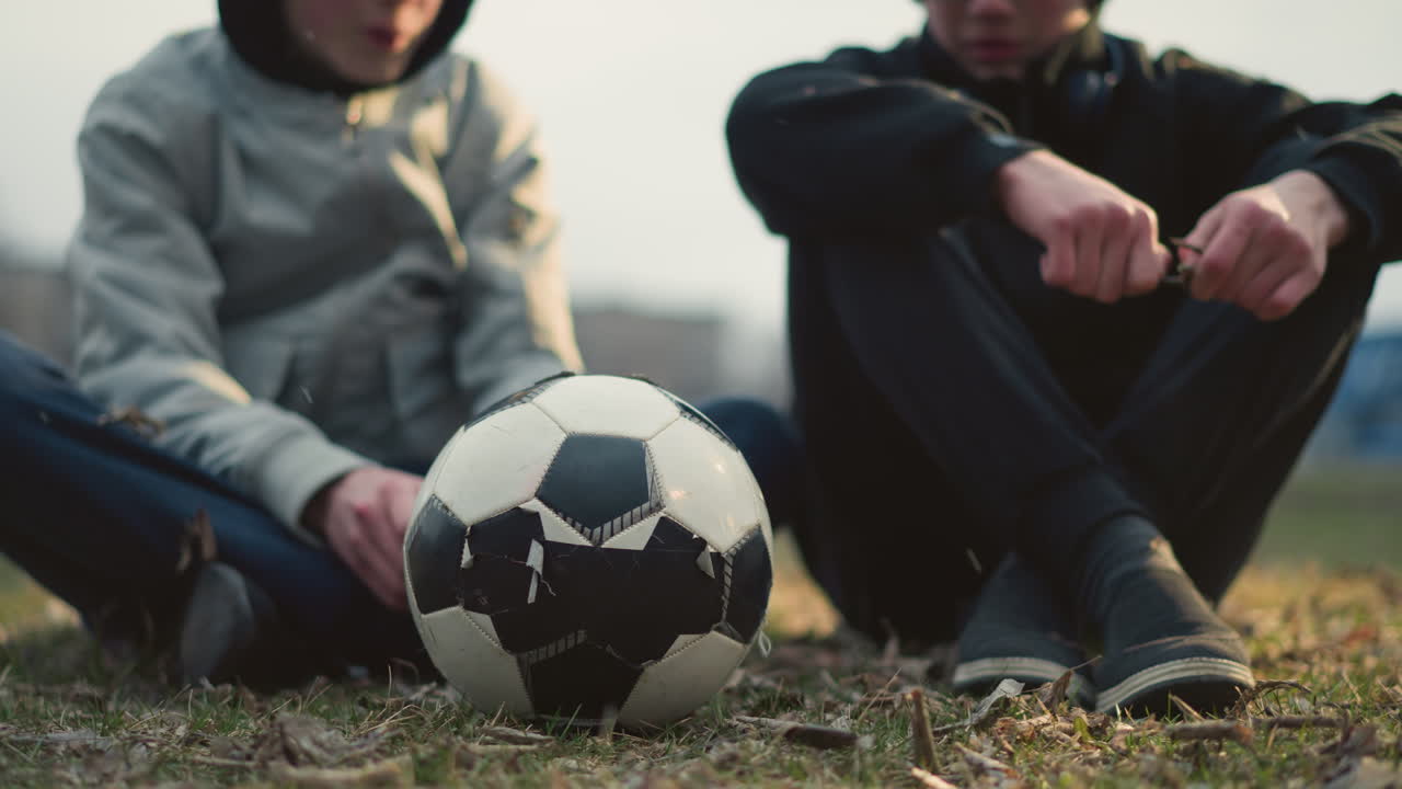 Blur view of two boys are seated on a grassy field, engaged in a discussion, the boy wearing glasses is holding a stick, attempting to cut it, the boy in a gray top has a soccer ball in front of him