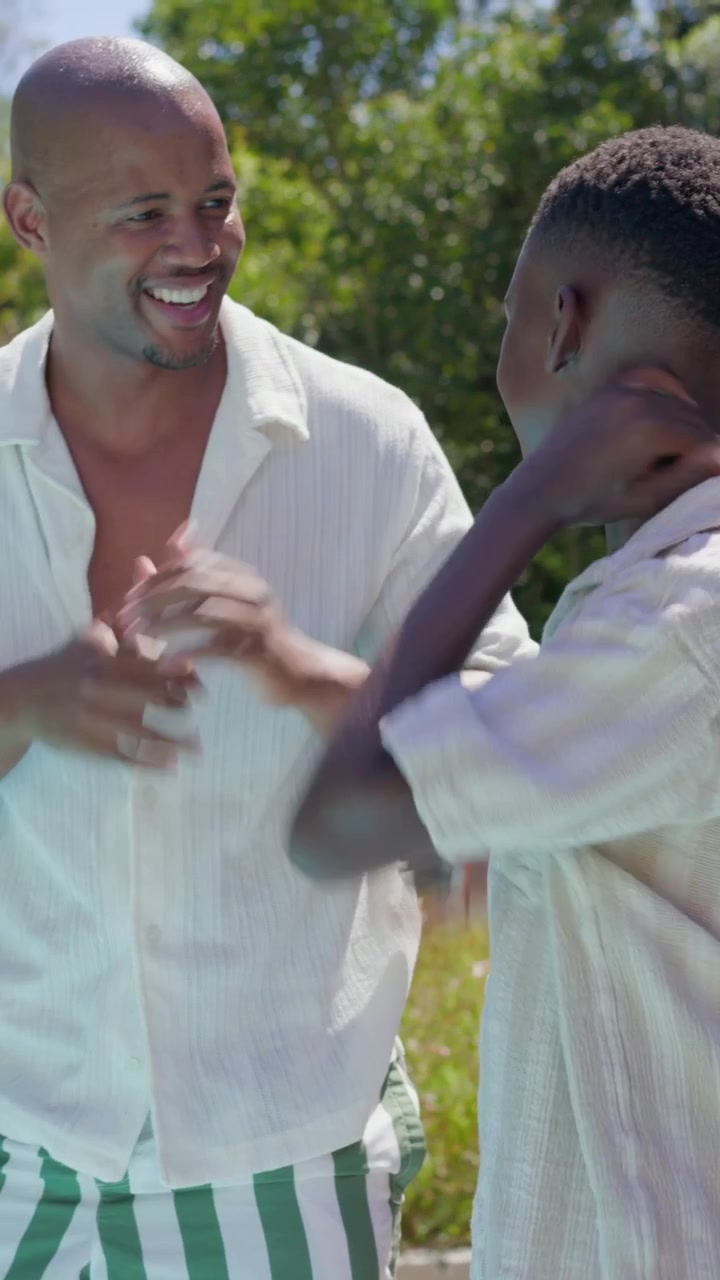 Video verticale: Papà sorridente che mostra un movimento del collo dopo che un giovane gli tocca il braccio sull'erba assolata, con camicia bianca.