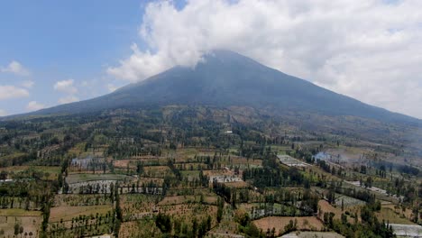 clouds flowing above sumbing mountain with agriculture fields bellow, aerial view