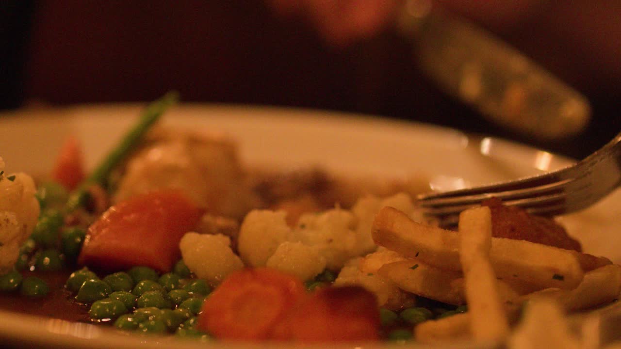 Diner uses fork and knife to eat steak pie with vegetables under warm, ambient lighting