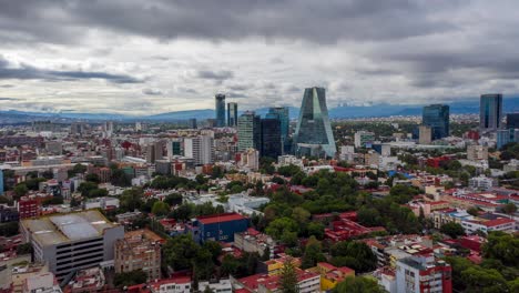 Hiperlapso:-Espectacular-Vuelo-Hacia-Atrás-Del-Centro-De-La-Ciudad-De-México-Sobre-Rascacielos,-Edificios,-Carreteras,-Carreteras-Y-El-Centro-Comercial-Plaza-Manacar-Con-La-Cordillera-De-Fondo-En-Un-Día-Nublado,-Drone