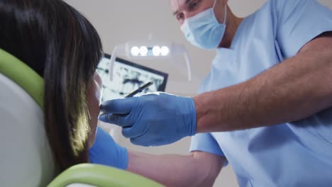 dentista caucásico con máscara facial examinando los dientes de una paciente femenina en una clínica dental moderna