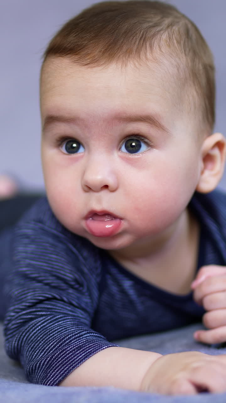 Lovely baby boy lies on belly looking up with surprise. Cute kid starts to move his limbs actively. Close up. Blurred grey backdrop. Vertical video