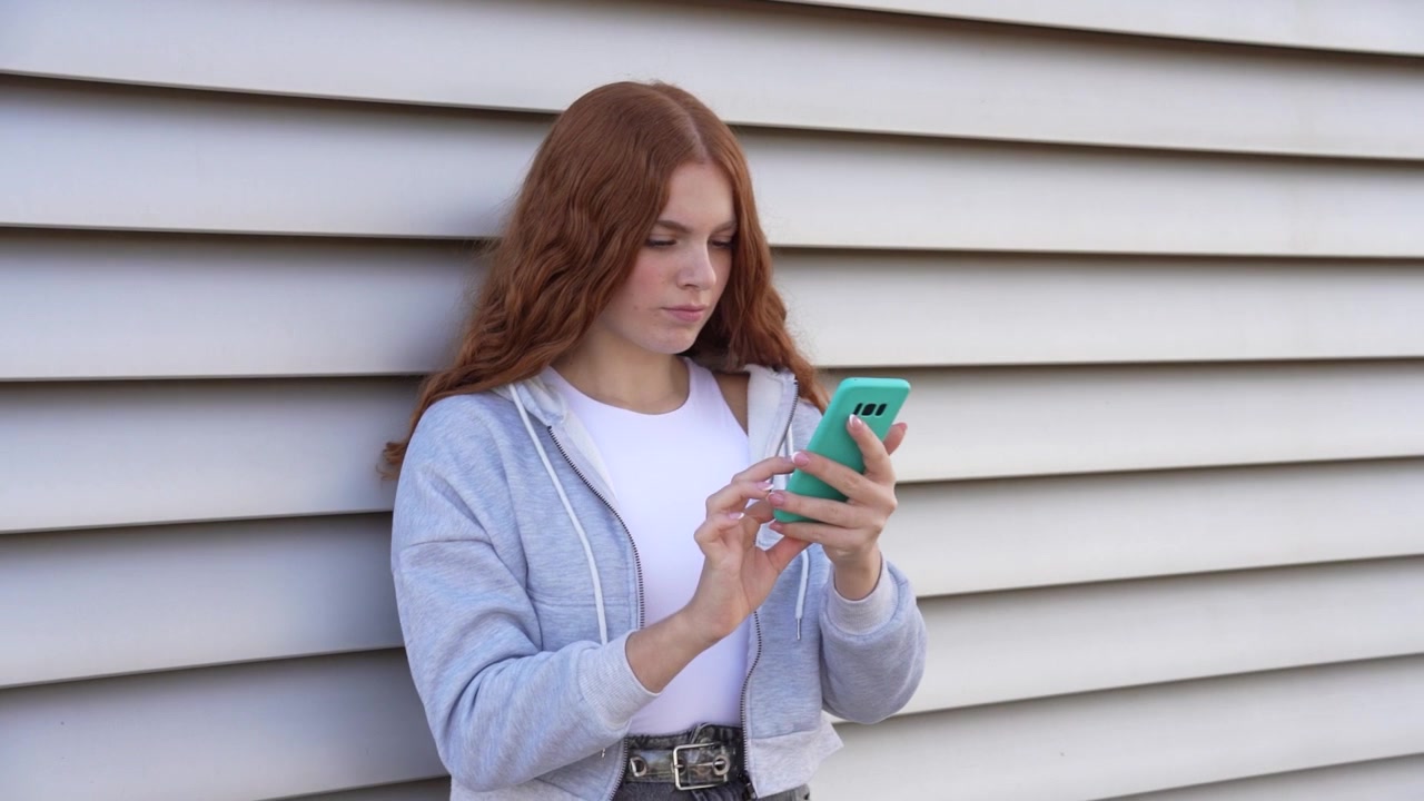 Una giovane donna è completamente assorta nel suo smartphone mentre è appoggiata casualmente a un muro.