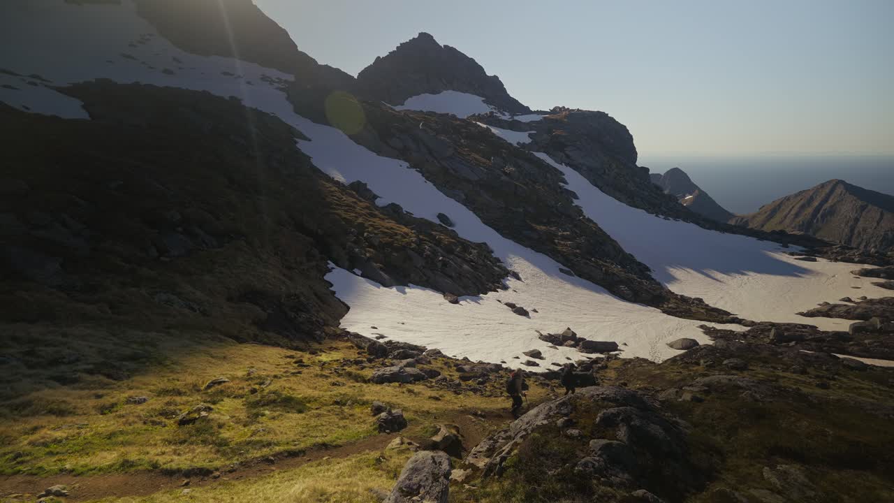 Vandrare går genom ett fantastiskt alpint landskap med snötäckta toppar, havsutsikt och mjukt ljus som lyser upp gräset i Lofoten, Norge. Statisk bild
