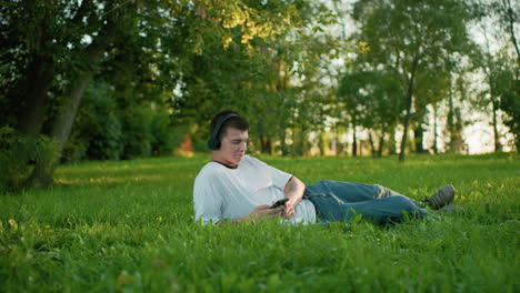 white man wearing headphones lying on grassy field holding smartphone, nodding to music, surrounded by vibrant greenery and trees with sunlight streaming through in background