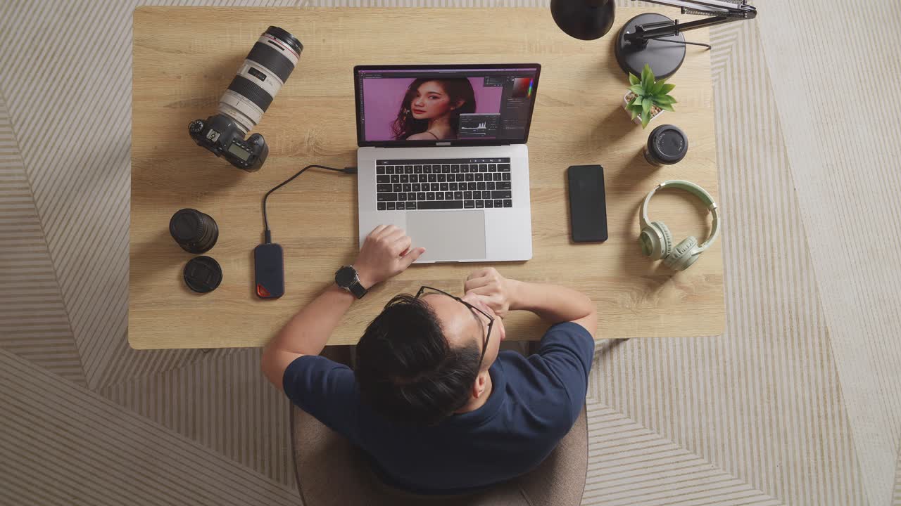 Top View Of Asian Male Editor Thinking About The Idea Then Raising Index Finger While Sitting In The Workspace Using A Laptop Next To The Camera Editing Photo Of A Female At Home