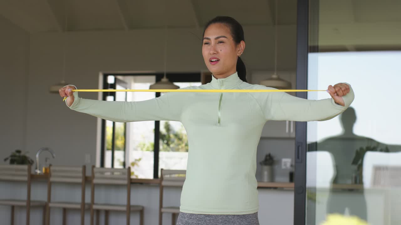 Asian woman spotting slack in yellow resistance band, pulling band outward at patio door