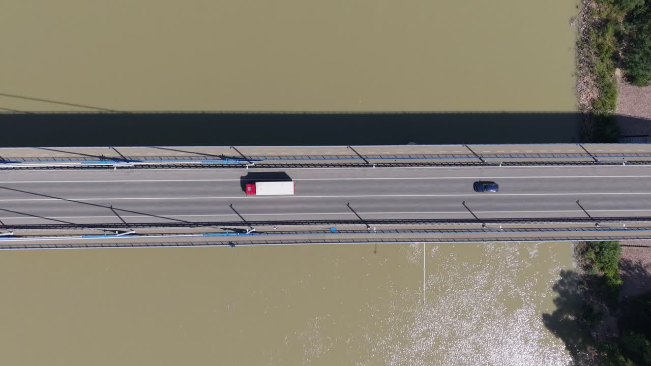 An aerial view of some truck and car crossing a suspension bridge over a muddy river. The scene captures the clean geometry of the bridge and the contrasting colors of the vehicles against the asphalt