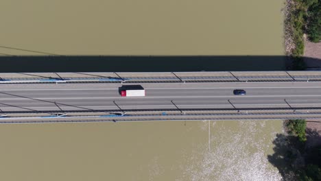 an aerial view of some truck and car crossing a suspension bridge over a muddy river. the scene captures the clean geometry of the bridge and the contrasting colors of the vehicles against the asphalt