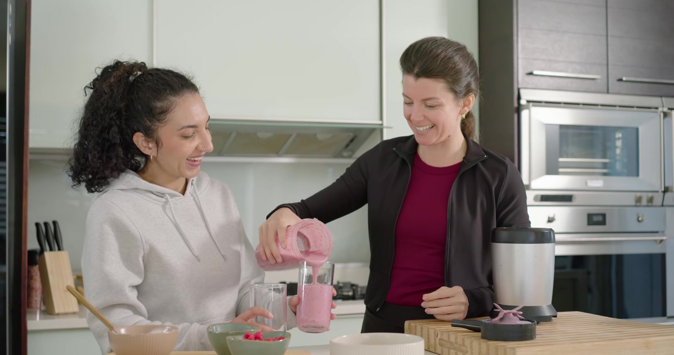 Il frullatore ha terminato; donne di diverse etnie sollevano una tazza e versano una bevanda rosa in altre tazze sull'isola della cucina per condividerla.