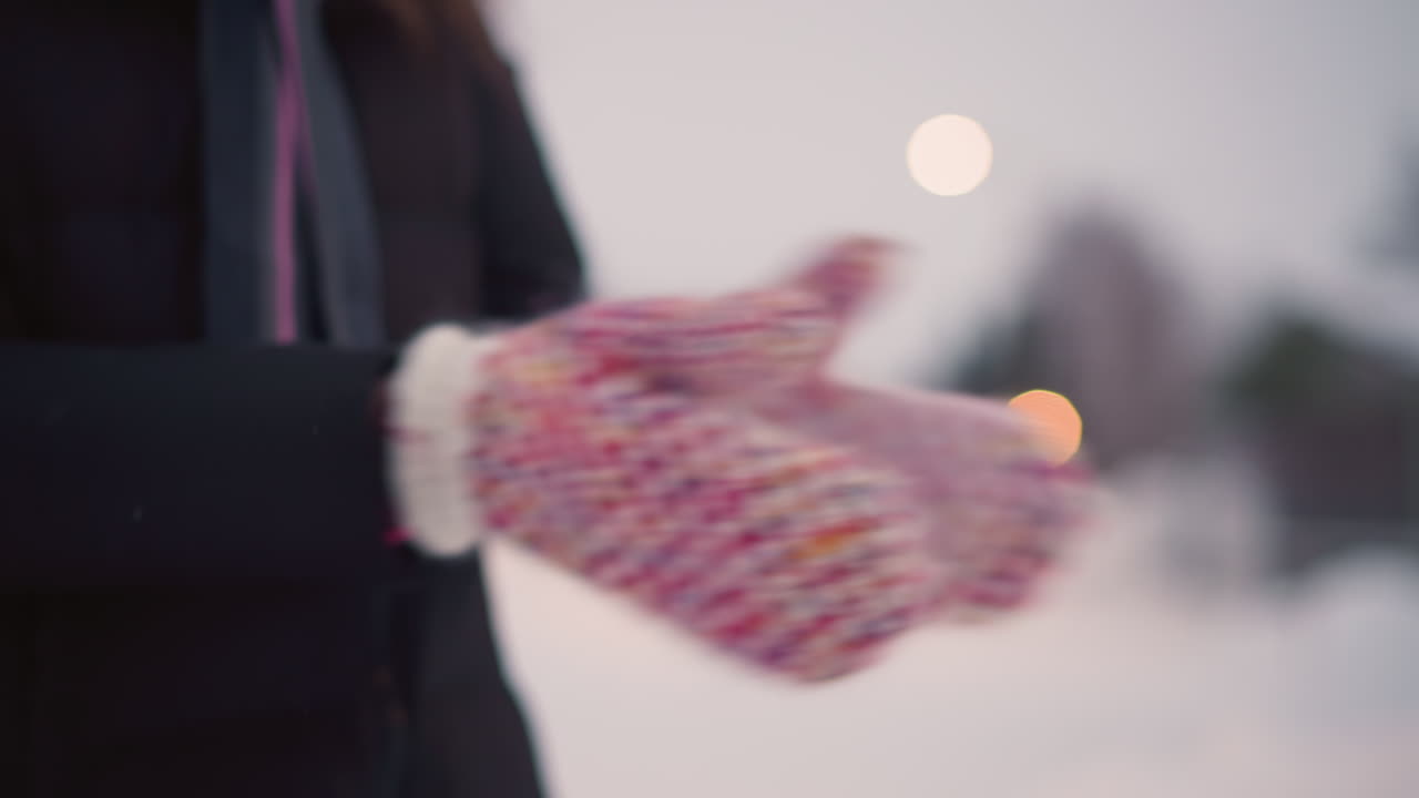 Skater wearing colorful knitted mittens claps hands in cold winter air, snowflakes scattering as warm breath rises, creating cozy seasonal atmosphere against soft glowing outdoor winter lights