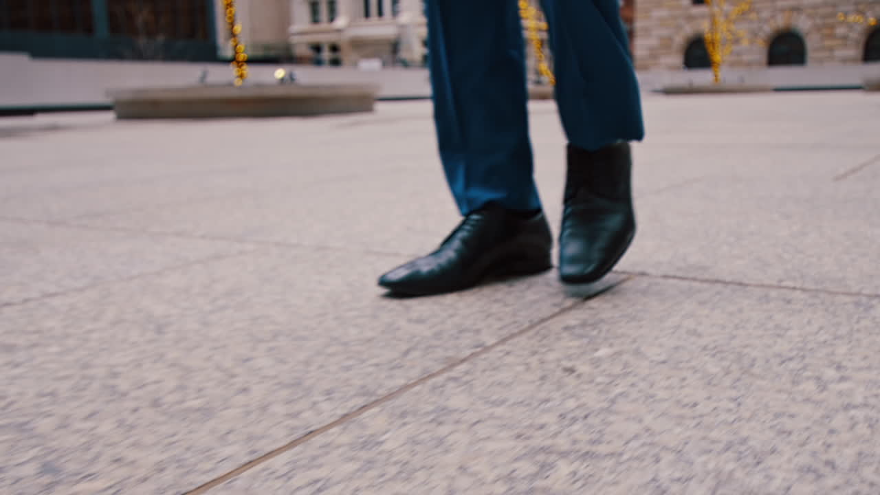 New York, USA, 28 May 2025: Close up of black leather shoes walking on city pavement. Low section shot of man in blue trousers and formal footwear walking on stone tiles