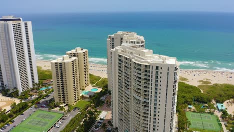 aerial drone shot slowly rotating around residential towers with the blue water of the atlantic ocean with waves crashing on the sandy beach as tourists sunbathe