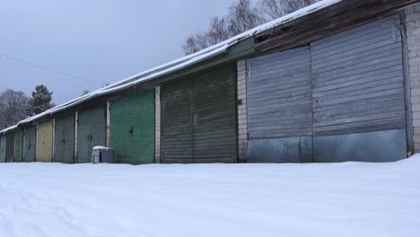 an old soviet styled garage complex made out of bricks that has different colored wooden doors or gates to them during winter time while thick white snow is covering the roof and ground. low angle.
