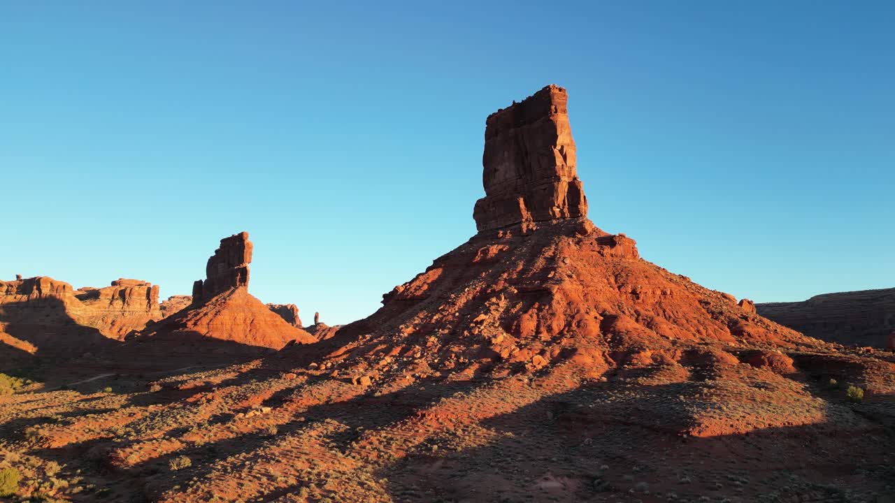Ripresa aerea con drone nella Valley of the Gods, Utah, USA, che mostra Castle Butte e Stagecoach Rock completamente illuminati dal sole basso del mattino. Movimento stabile sopra le formazioni di arenaria rossa sotto un cielo azzurro limpido.