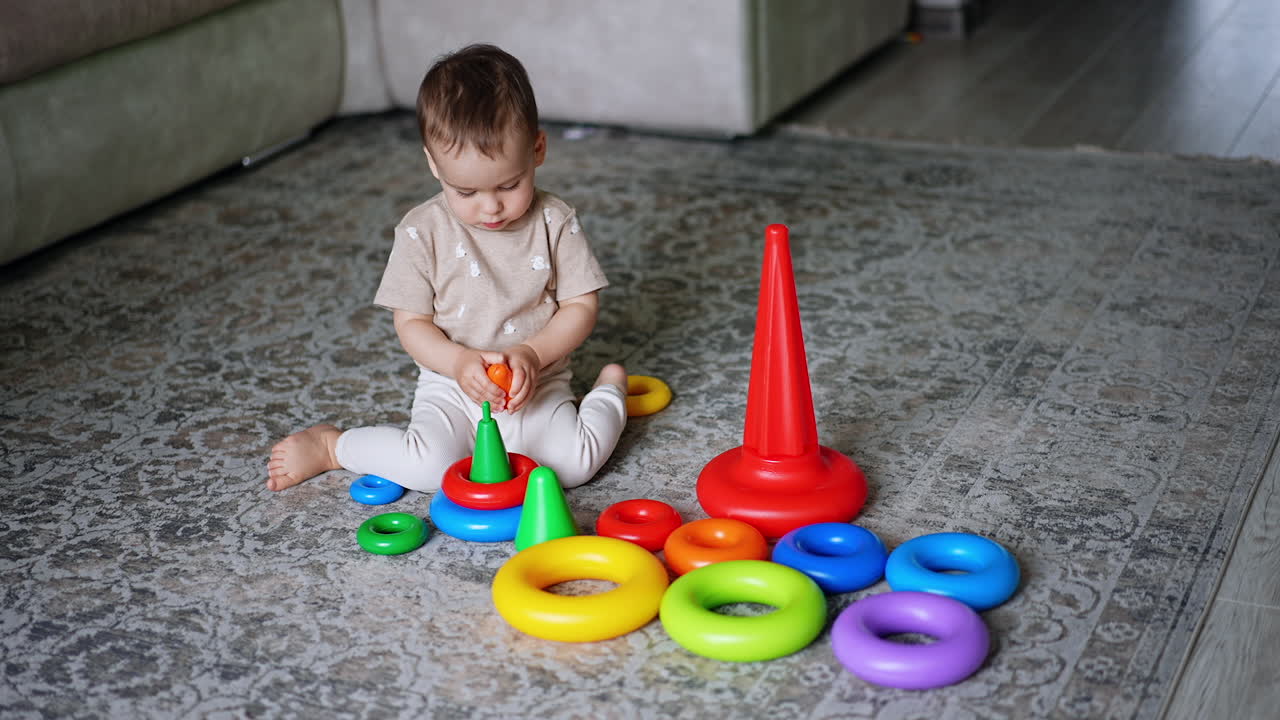 Cute toddler sitting on the floor holding a piece from toy pyramid. Baby boy notices something, stands up to go there.
