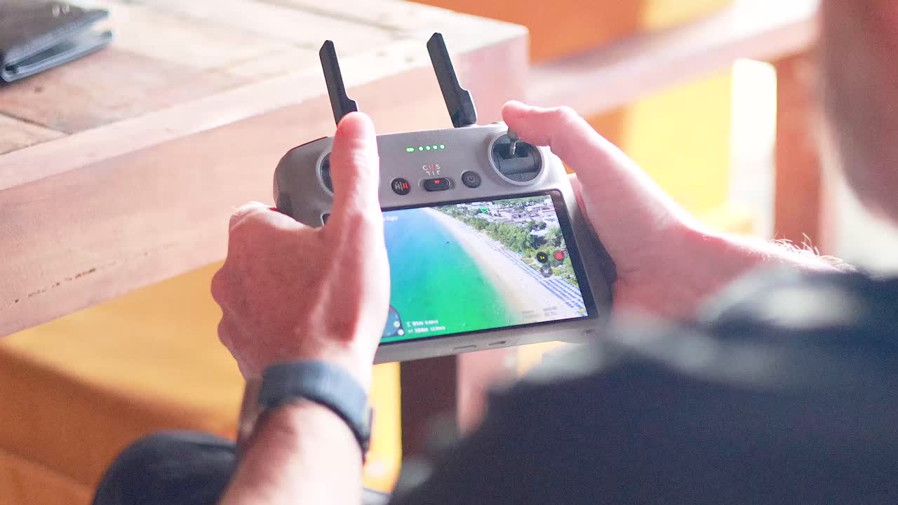 Hands operate a drone controller, displaying a live aerial view of a Phuket beach. Bright lighting enhances the scene