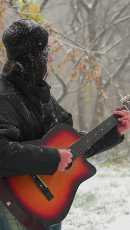 vista lateral de una mujer con capucha negra tocando guitarra acústica al aire libre en un parque nevado, rodeada de árboles desnudos helados y nieve que cae suavemente, creando un momento de invierno sereno lleno de música
