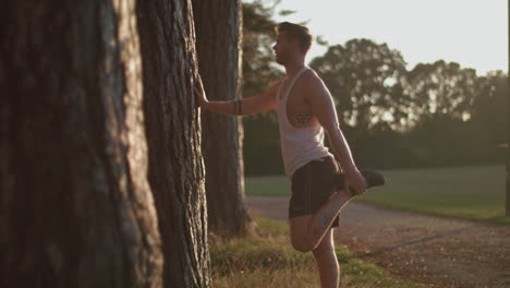 Hombre-En-El-Parque-Estirando-Las-Piernas-Antes-De-Salir-A-Correr-Mientras-El-Sol-Se-Pone-Detrás-De-él-En-Cámara-Lenta