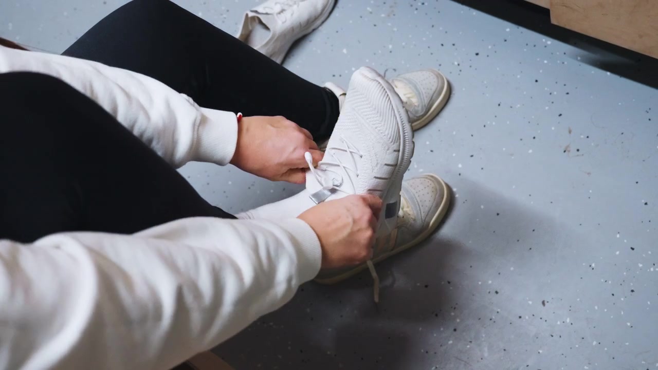 Mani in primo piano che allacciano i lacci delle scarpe da ginnastica, preparazione tranquilla su una panca, felpa bianca e leggings neri, pavimento interno testurizzato, movimenti delle dita precisi che assicurano il nodo, prontezza atletica concentrata.