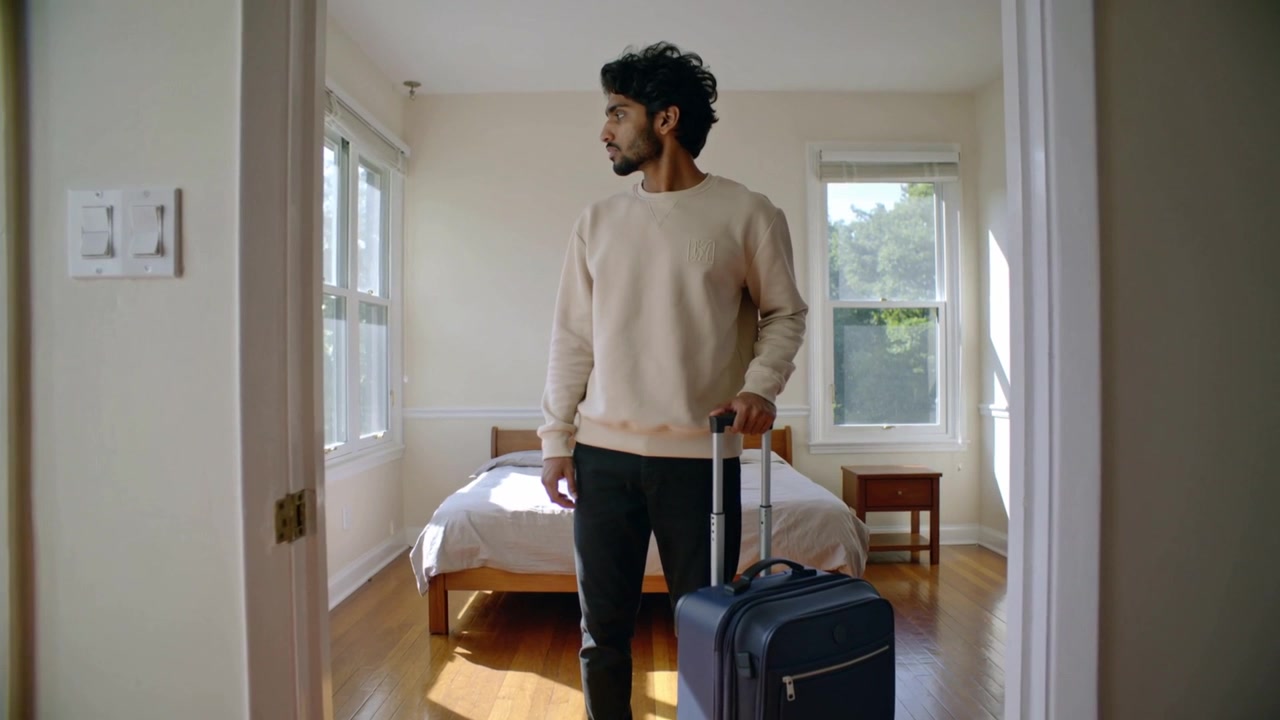 Entering adult man in light sweater and dark trousers holding suitcase and scanning bedroom windows