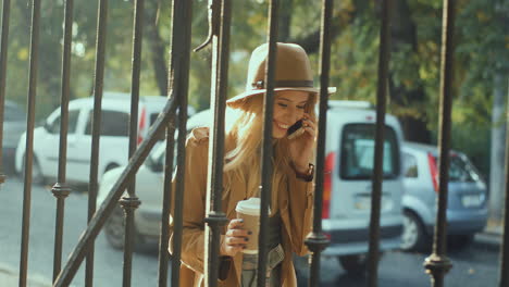 alegre joven rubia sonriente con un sombrero y un abrigo caminando detrás de una cerca, bebiendo café para ir y hablando por teléfono en la calle