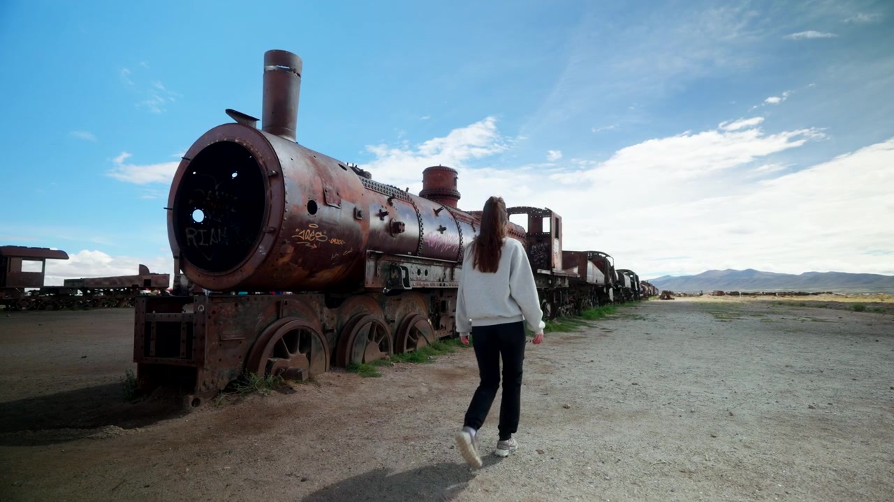 Jovem mulher caminha ao lado de uma locomotiva a vapor enferrujada no famoso cemitério de trens em Uyuni, Bolívia. Ideal para conceitos de viagem, aventura, patrimônio e indústria abandonada
