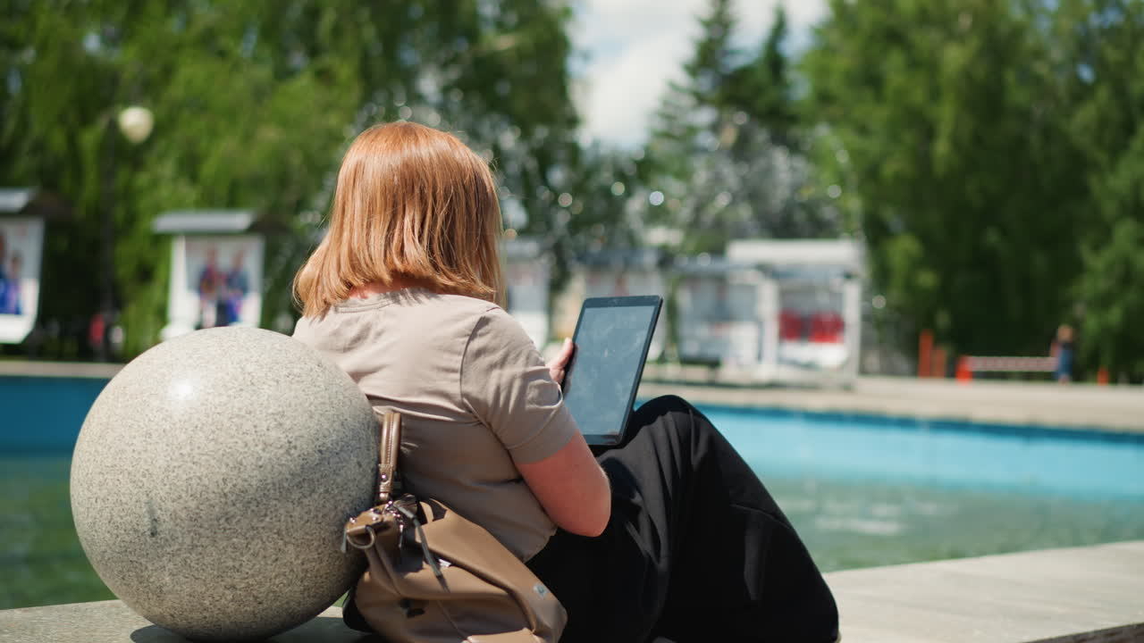 Young lady sitting relaxed by city fountain using tablet and smiling under bright summer sunlight, brown bag resting beside her, calm mood and casual posture capturing harmony of leisure