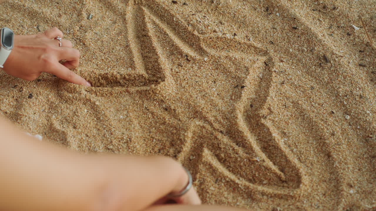 Händer ovanifrån ritar en stjärna i sanden, ung kvinna med glasögon och klocka skissar en femuddig form över den korniga stranden, lekfull sommar energi, svepande fingerlinjer, texturerad detalj, nostalgisk semester.