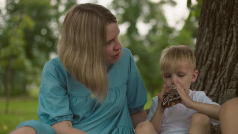 a mother watches her young son attentively as he eats his snack, while a partial view of another person can be seen nearby