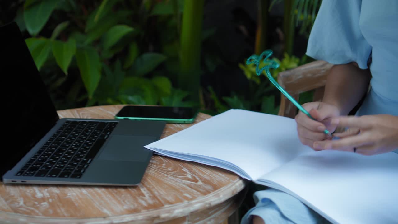 푸른 드레스를 입은 젊은 유럽 소녀가 노트북에서 원격으로 온라인으로 작업하고 초록색 식물과 함께 뒷마당의 화면을보고 있습니다.